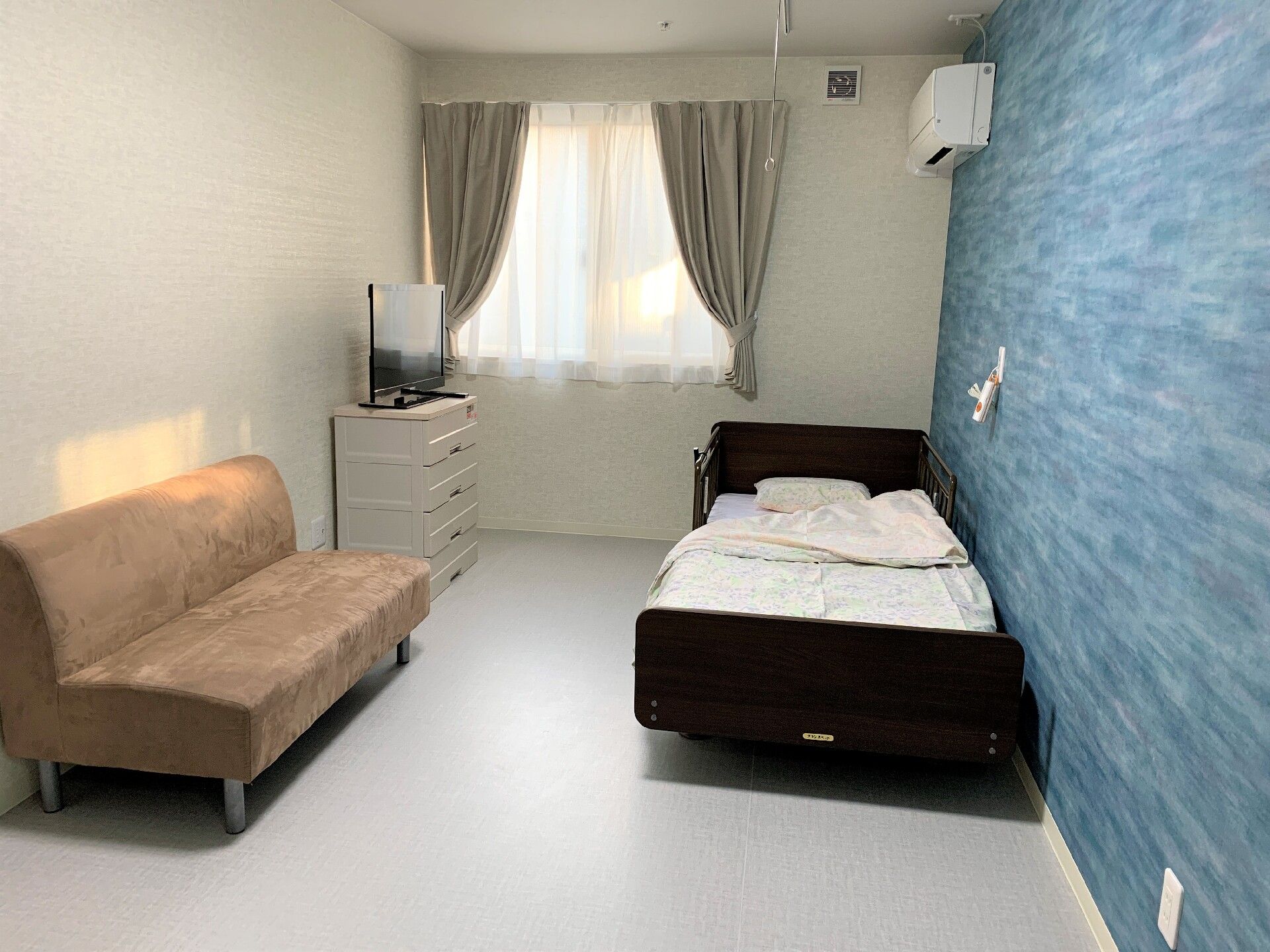 ベッドのそばにソファが置かれている施設内の居室。エアコンとテレビ、ベッドが置いてある空間
