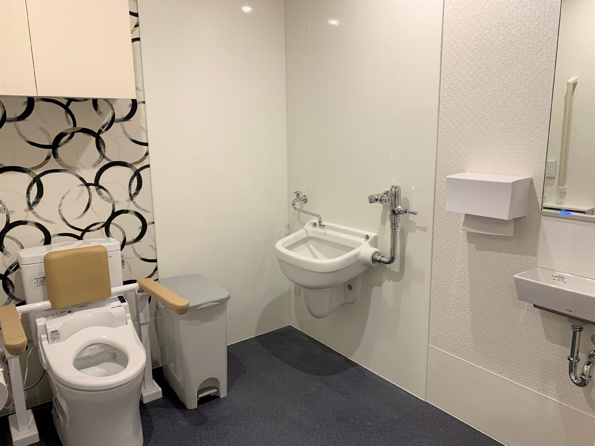 手すりがついたモダンな壁紙のトイレと洗面台がある施設内のトイレの様子。