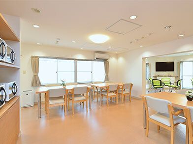 大きな窓が二つ並んで設置された食堂には電子レンジが複数置かれ、木造のダイニングテーブルセットやポップなデザインのテーブルセットがあり、テレビも置かれている。