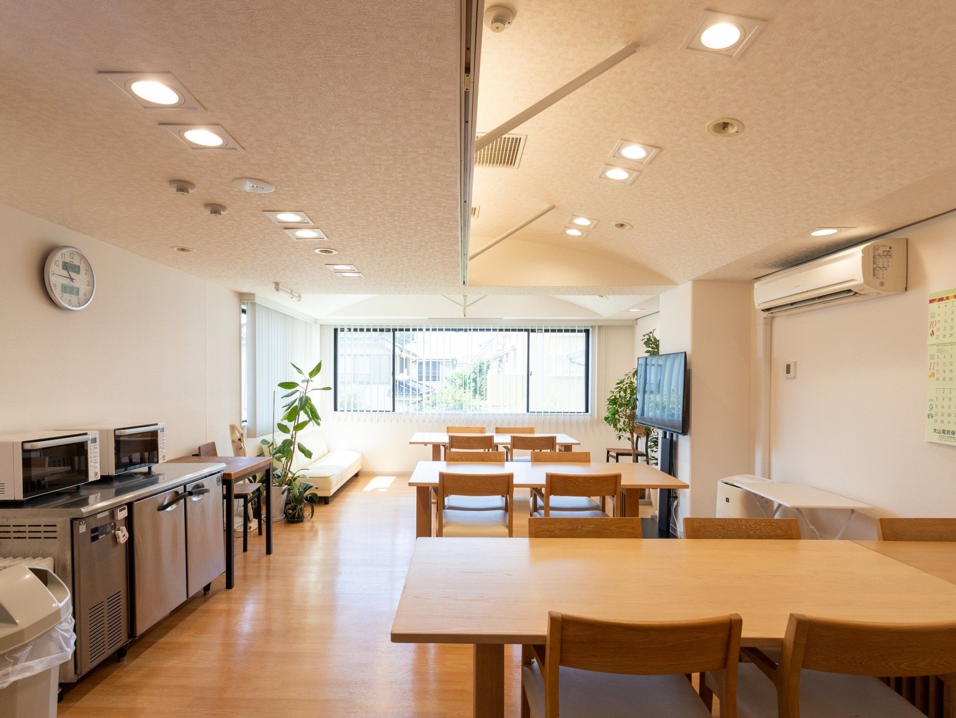 木製のダイニングテーブルセットが複数置かれた明るい食堂には電子レンジがたくさん設置され、テレビの前にはデザインの異なるテーブルセットがある。
