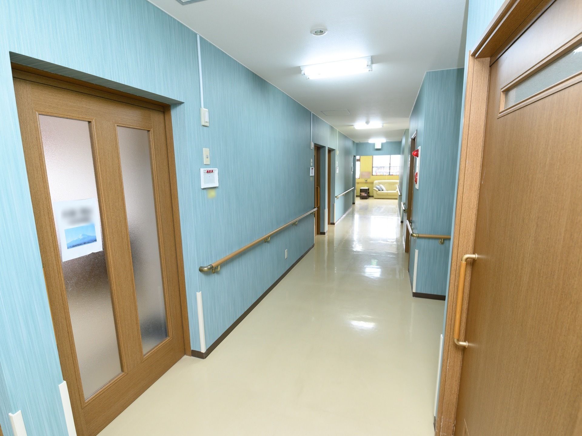 青色の壁になっている廊下がある。居室などの入口以外の部分の壁には手すりが設置されている。非常ベルがついている。