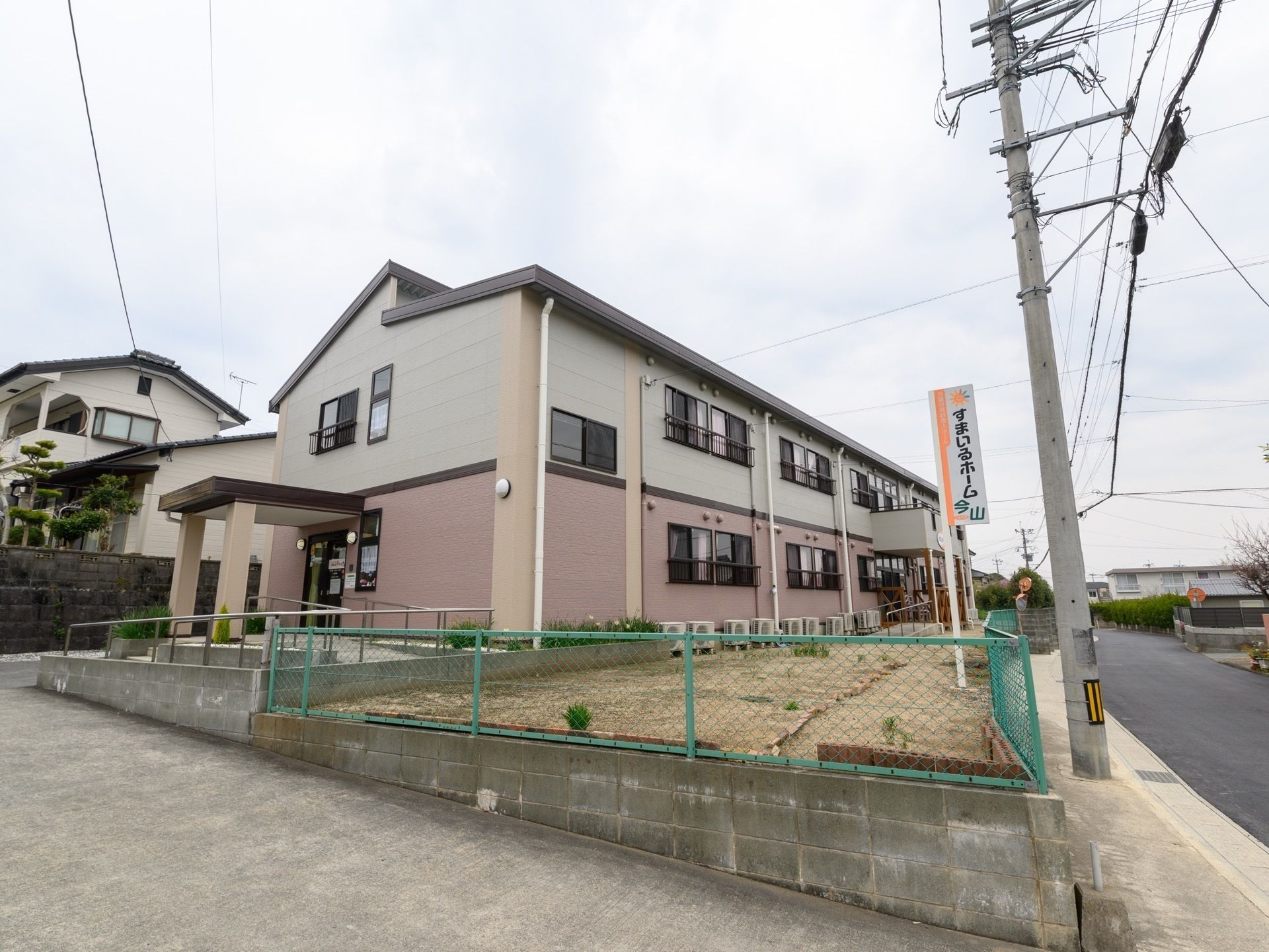 道路から少し高くなった敷地に建つ２階建ての施設である。道路沿いにフェンスの中に、施設名称が縦に書かれた看板が設置されている。