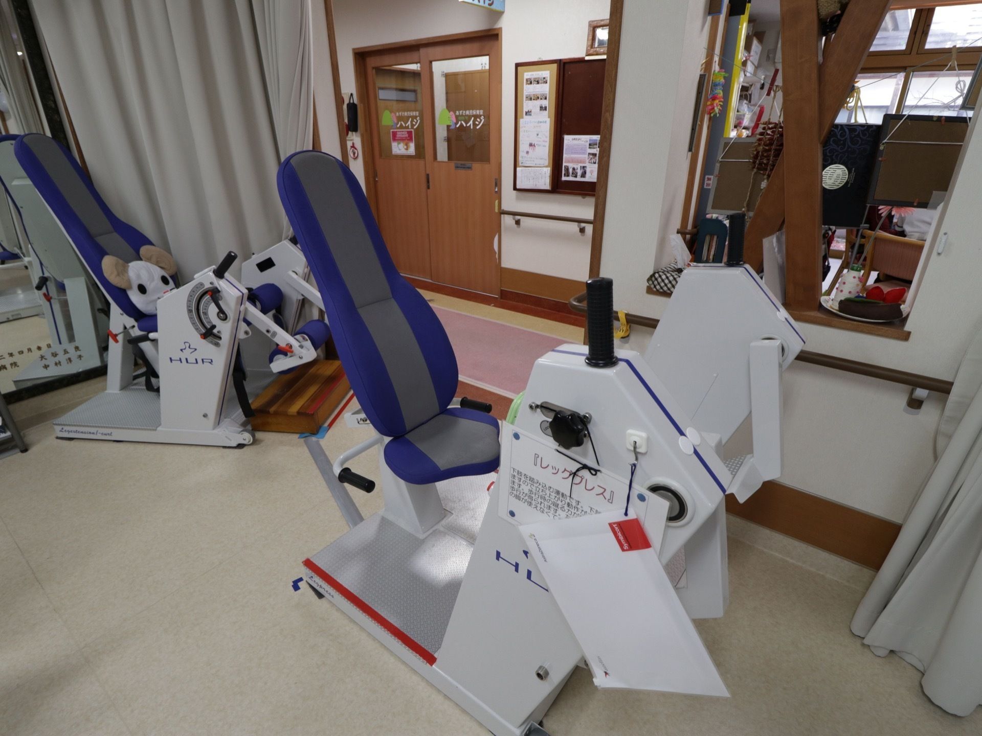 足の動きを鍛えるマシーンが２台置かれている。手すりが壁に取りつけられている。床はカーペットになっている。