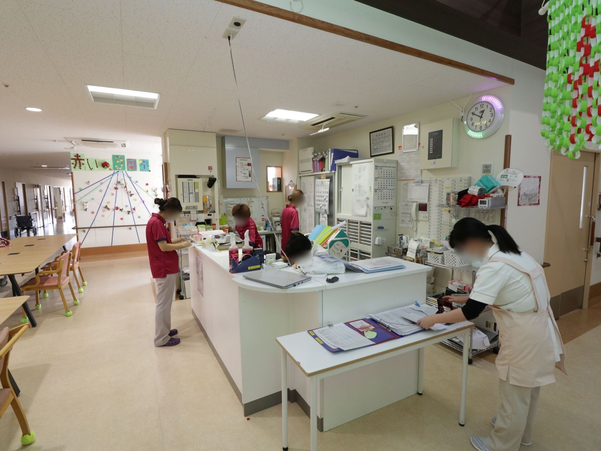 書類や電話などが置かれており、スタッフがそれぞれ異なる作業を行っている。壁には時計が掛けられている。