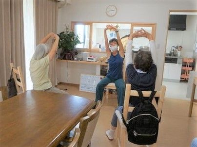 手を挙げて体をほぐしている高齢者とそれを指導するスタッフの様子