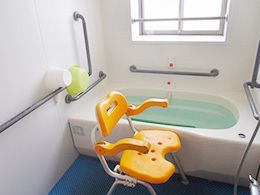 オレンジの呼び出しボタンやグレーの手すりが付いた白を基調とした浴室。一人用の浴槽があり、シャワーチェアもある。