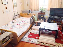 花柄の寝具がセットされたベッドが置かれた部屋にはテレビやローテーブル、扇風機などがあり、にぎやかな雰囲気。