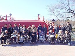 車いすの高齢者たちと家族、スタッフが一緒に外出を楽しんでいる。大きな赤い橋の前で記念撮影。