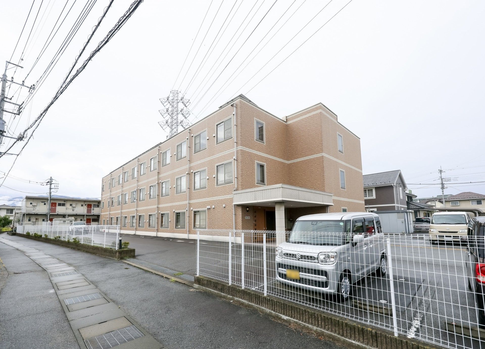 ブラウン系の建物は3階建てで周りには建物はなく、玄関には屋根があり、駐車場も完備されている。