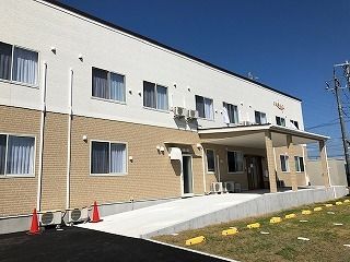 ベージュと白の2階建ての建物はフロアにより外壁の色が異なり、芝生が敷かれた駐車スペースが前にある。