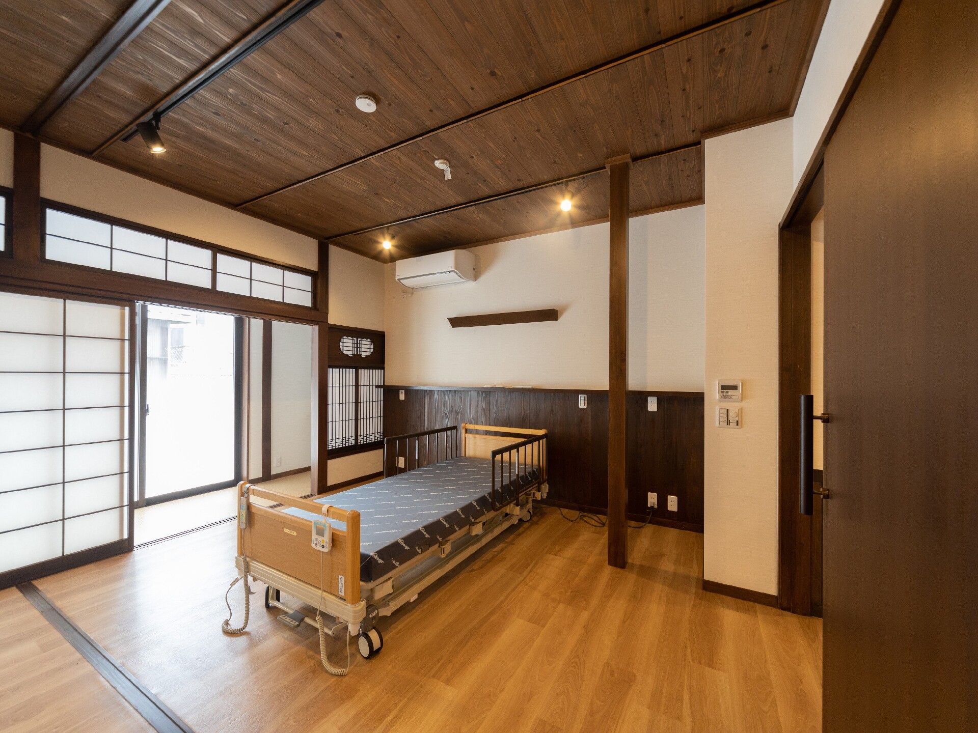 エアコンが付いたフローリングの部屋は天井が高く、介護ベッドが置かれ、右側には木製のスライド式のドア、左には障子が設置されている。