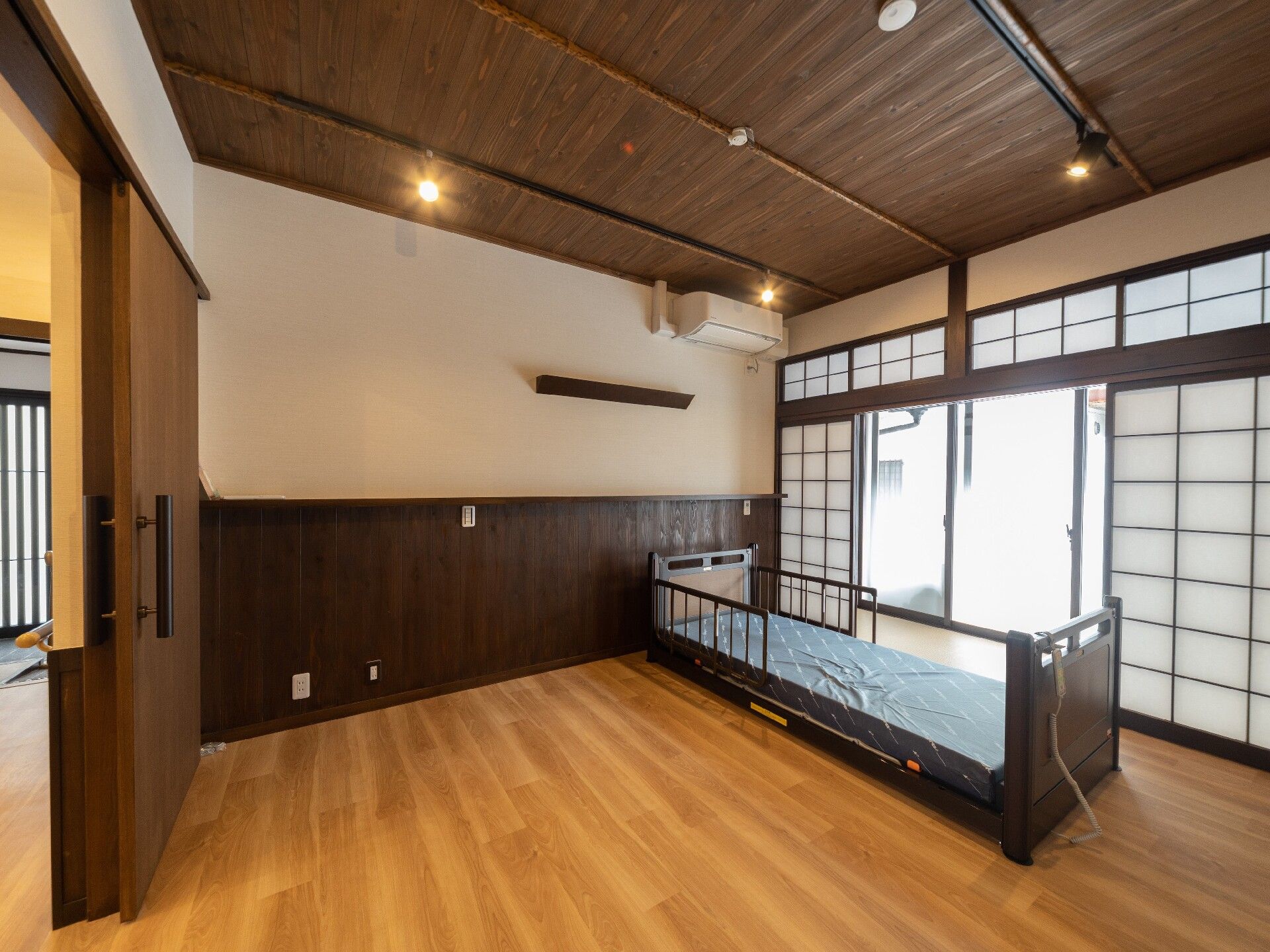 暖色系の色の間接照明がたくさんつけられた部屋はフローリングで壁にはエアコンが付き、中央に介護ベッドが置かれ、窓には障子がある。