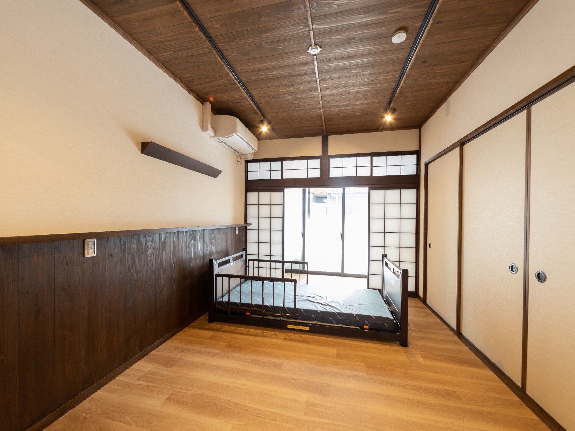 障子が設置されたフローリングの部屋にはエアコンや介護ベッドがあり、右側の壁にはふすまが付いている。テレビや寝具、タンスなどの家具はない。