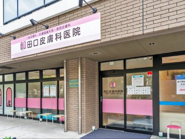 ブラウンのレンガ調の外壁の皮膚科医院。大きな窓がたくさん付き、外には待つ人のための椅子がある。