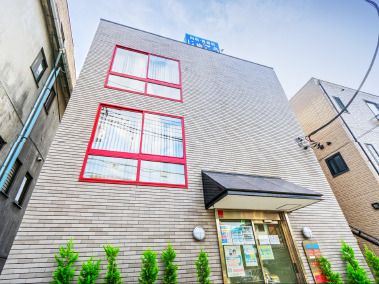 赤い枠の大きな窓が付いた3階建ての建物はブラウン系の外観で玄関の周りには草木が植えられている。