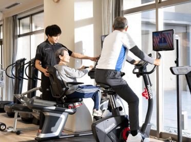 黒い上下に身を包んだ男性トレーナーが笑顔でトレーニング中の眼鏡をかけた高齢女性のそばにいて声かけをしている。