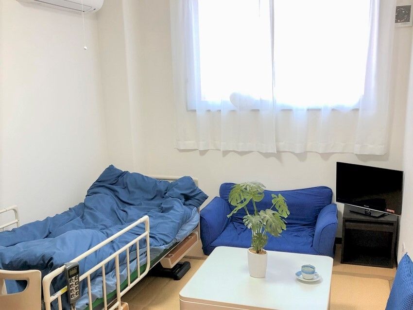 テレビやソファ、寝具付きのベッド、テーブルなどが置かれた部屋。壁にはエアコンが付き、窓にはカーテンが付いている。