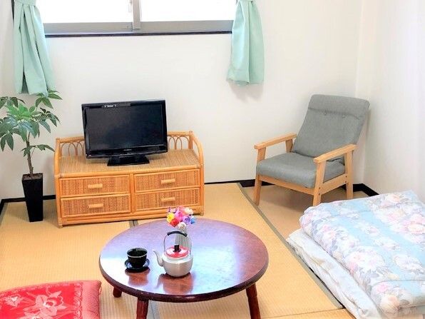 小さなやかんや湯飲みが置かれた円形のちゃぶ台やテレビ、観葉植物などが畳の上に置かれ、敷布団もたたまれた状態で置かれている。