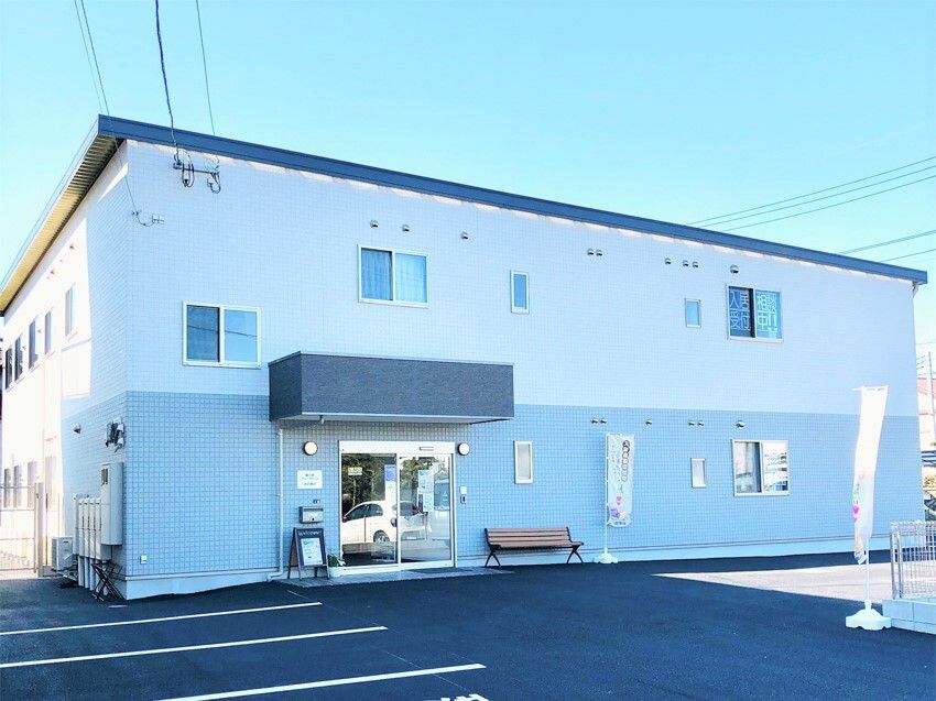 2階建ての建物は1階部分がグレー、2階部分が白い外壁で、玄関の横にはベンチがあり、広い駐車場も付いている。日当たりも良好。