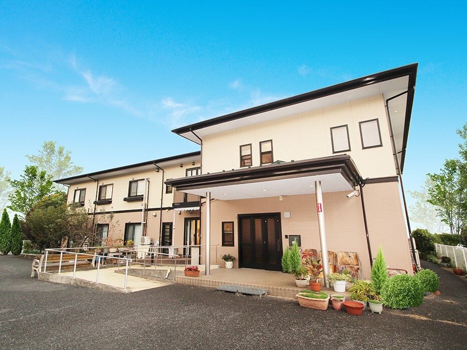 2階建ての建物はベージュ系で敷地内にはたくさんの木や花が植えられ、玄関周りには手すり付きのスロープもある。