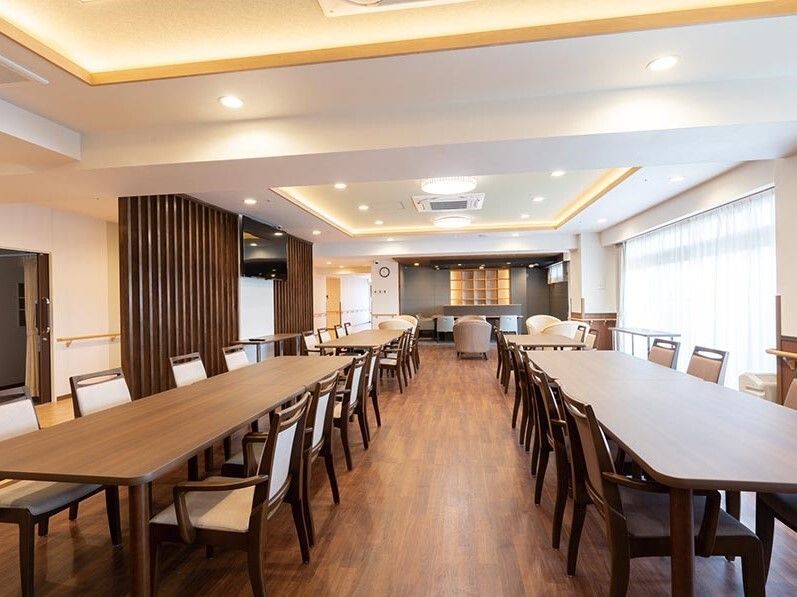 木製の大きなダイニングテーブルが複数置かれた食堂。奥にはリビングもあり、大きな窓も付いている。
