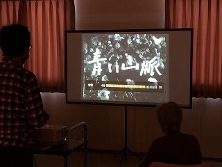 「青い山脈」と書かれたスクリーンを見ている高齢者。室内は明かりが消え、カーテンが閉められていて暗い。