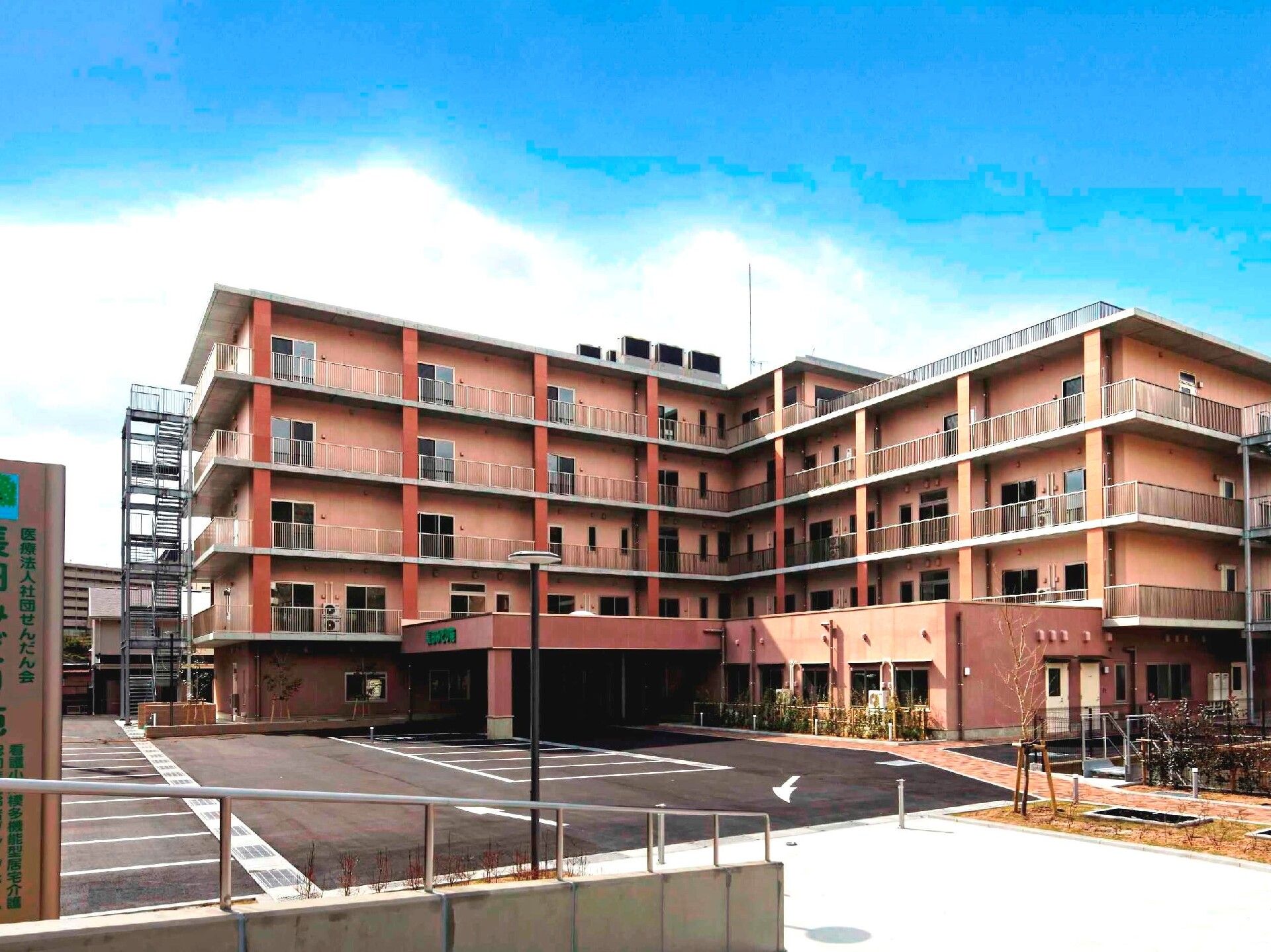 髪を後ろで一つに束ねた若い女性スタッフが、黒いニット帽をかぶってグリーンのチェックの車いすに座った高齢女性を抱きかかえる様にして介助している。