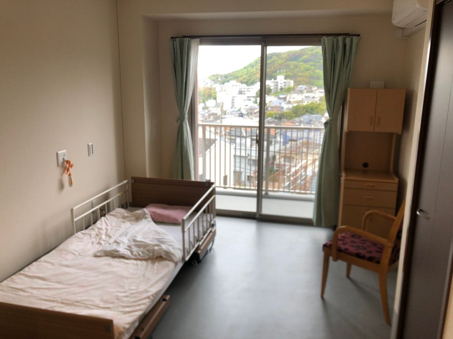 ベッドサイドには緊急通報装置が付き、収納家具やいす、寝具付きのベッドが置かれたシンプルな居室。大きな窓にはカーテンも設置されている。