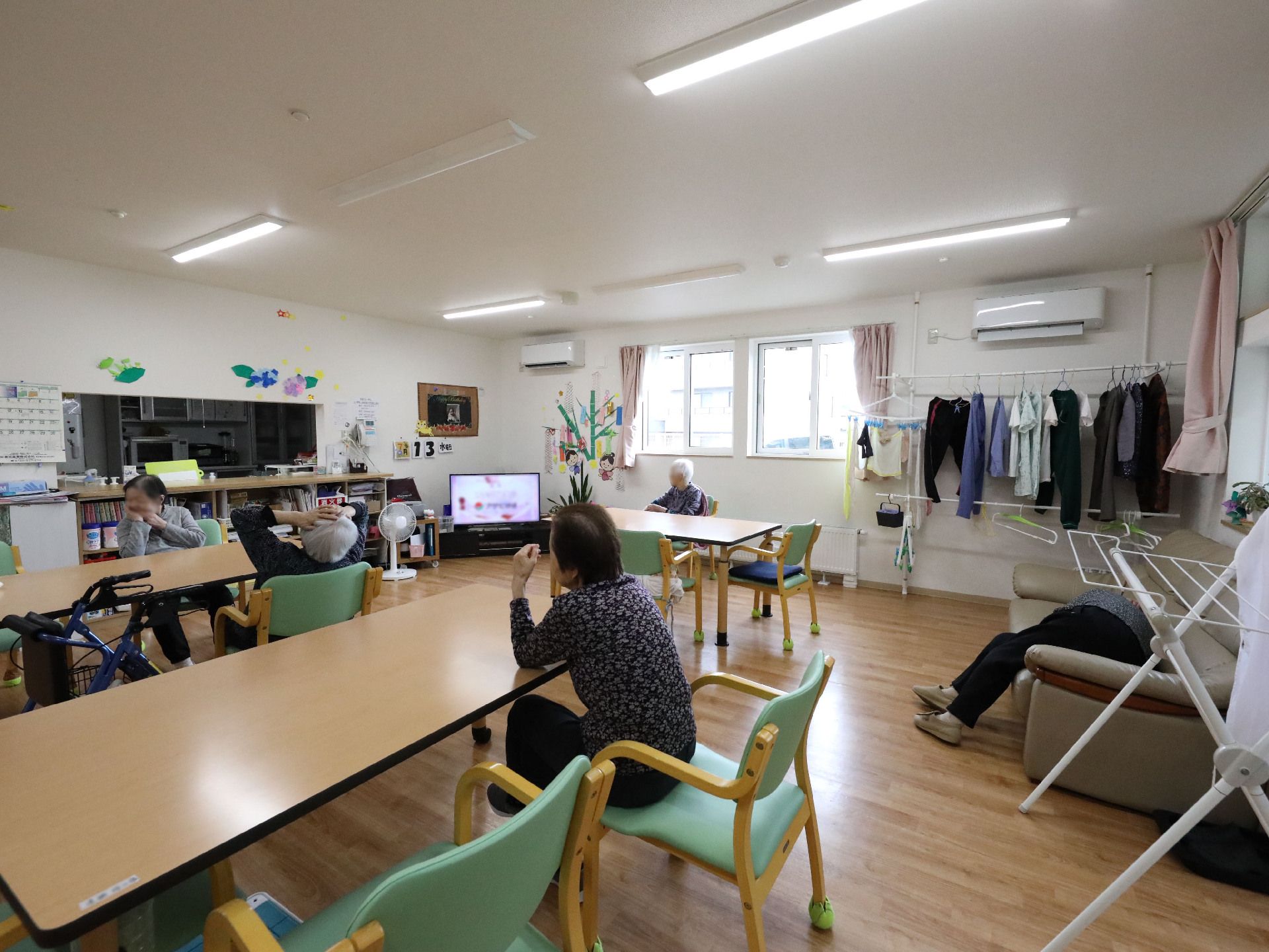 窓のある大きな部屋に並んだ椅子に座ってテレビを見る入居者様