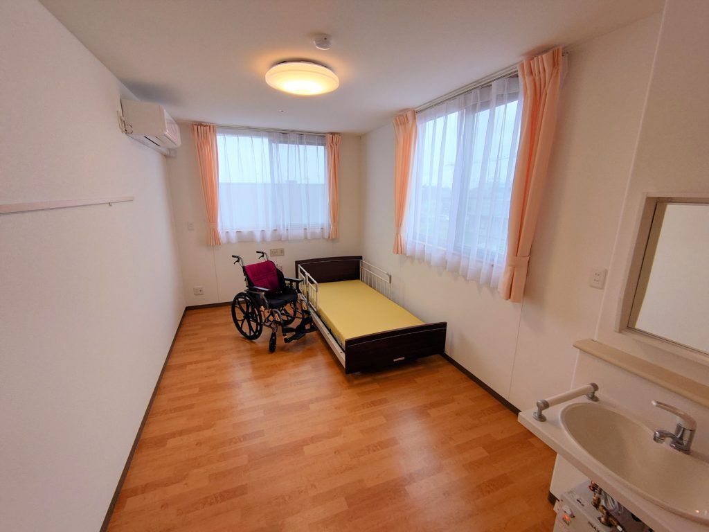 レースカーテンとカーテンが付いた大きな窓が２つあるフローリングの部屋には介護ベッドや車いすがあり、エアコンも付いている。