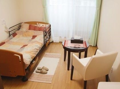 ベッドサイドには緊急通報装置が付いたフローリングの居室。大きな窓にはカーテンとレースカーテンが付き、いすとテーブル、テーブルの上にはティッシュがある。