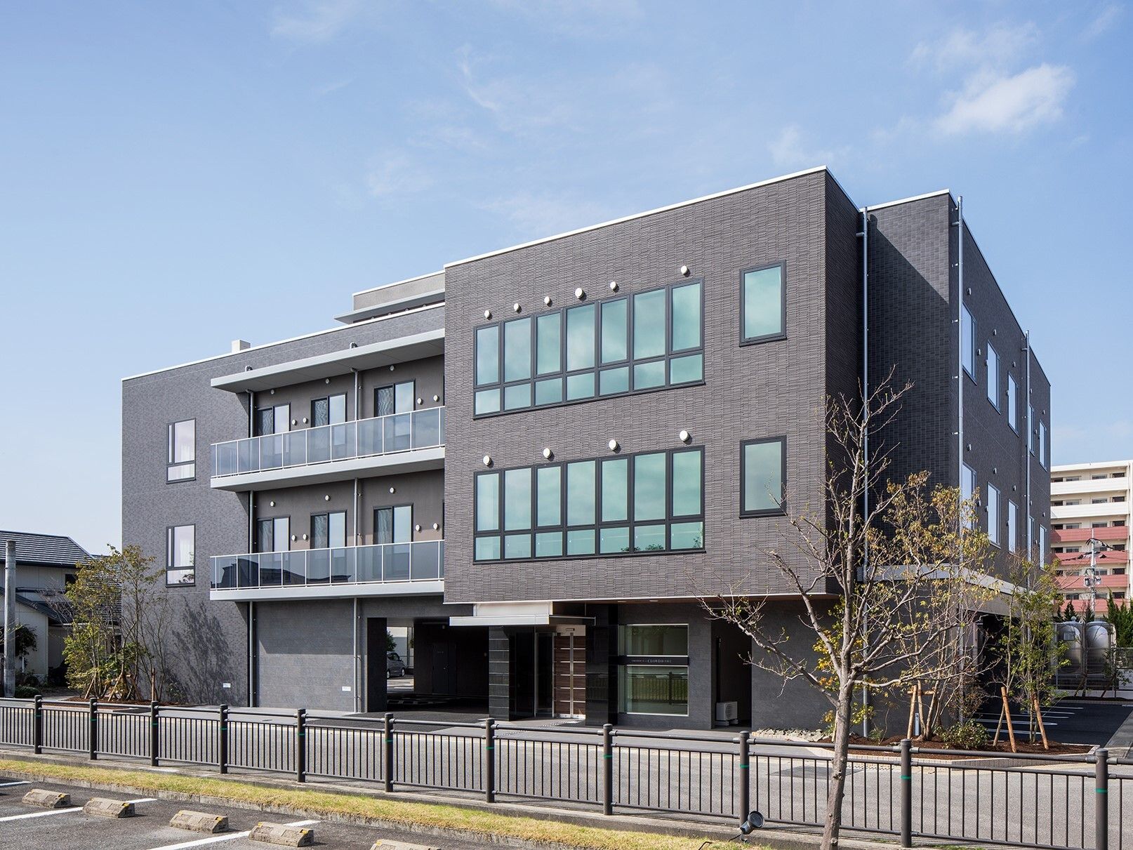 3階建ての建物はグレー系でスタイリッシュなデザイン。敷地内には駐車場があり、周囲には黒い柵もたてられている。周りには建物があるが日当たりも良好。