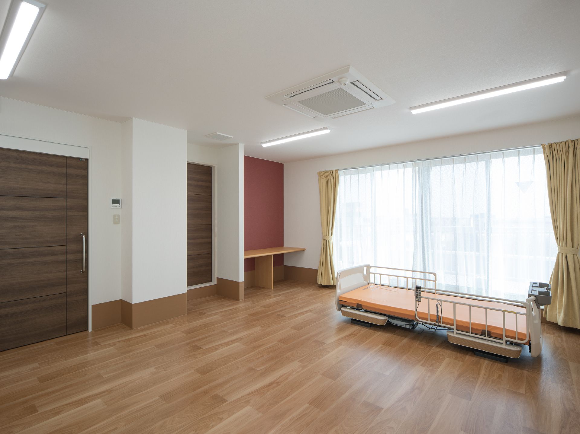 寝具がセットされた介護ベッドが設置された広い部屋にはグレーのソファの応接セットがあり、大きなテレビが窓のそばにある。