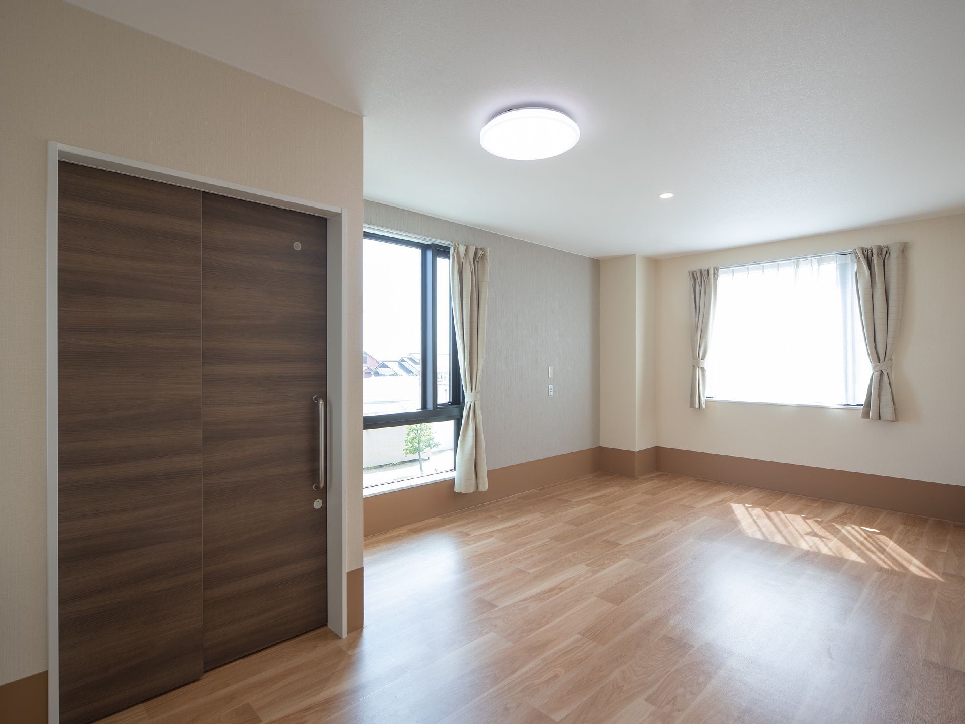 大きな窓が二つ付いたフローリングの部屋。窓にはカーテンが付き、照明が天井にあるだけで、家具や家電は置かれていない。