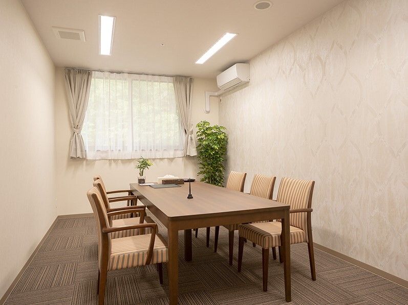 観葉植物が置かれたカーペット敷きの部屋には6人掛けのテーブルセットが置かれ、カーテン付きの窓もある。
