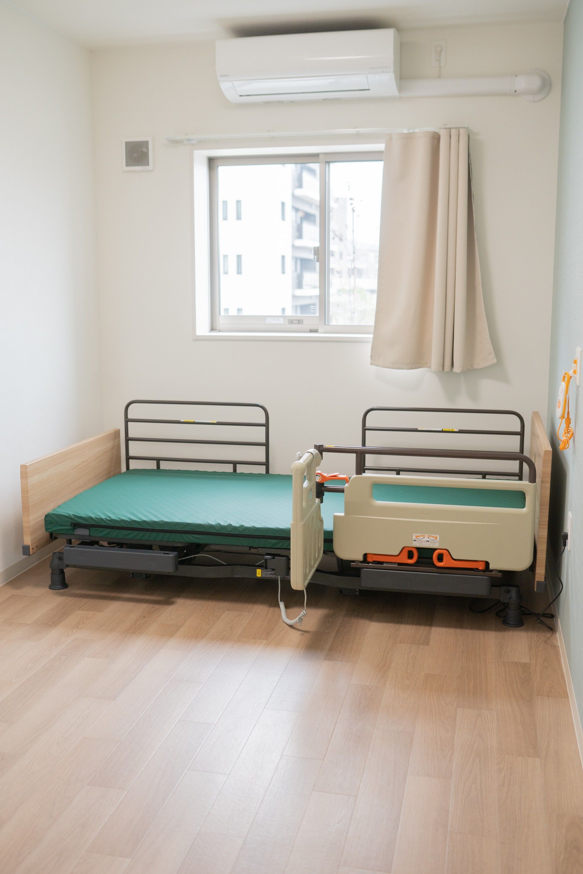 介護ベッドには白い寝具がセットされ、窓にはベージュのカーテンが付き、白い壁にはエアコンが設置されている。床はフローリング。