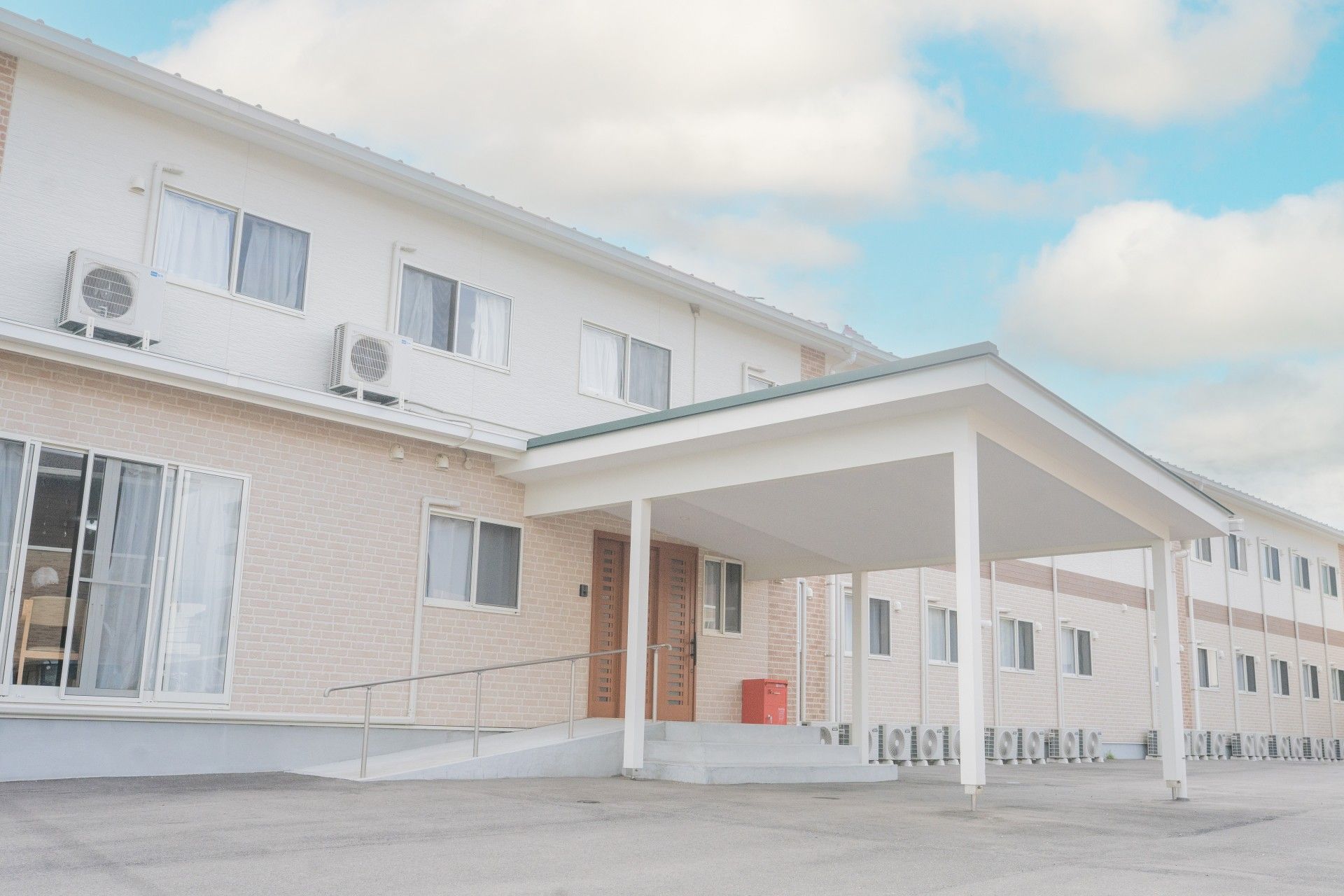 ベージュの2階建ての建物は窓がたくさんつき、玄関には手すり付きのスロープと屋根が付き、建物の周囲には背の低い緑の植物が植えられている。