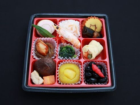 赤と黒の弁当箱は細かく仕切られ、それぞれの枠の中に一品ずつ入っている。筑前煮や栗きんとん、黒豆、昆布巻き、かまぼこなど正月風の献立。