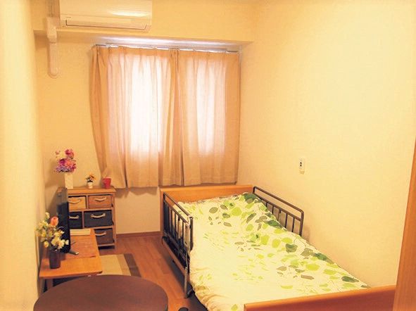 カーテンが付いた大きな窓があるフローリングの部屋にはエアコンや寝具付きのベッド、テーブルなどを置いている。