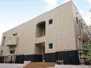 2階建ての建物は黒いフェンスで囲まれ、玄関には階段が付き、敷地内には緑も植えられている。空には青空が広がっている。