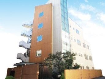 ブラウンと白の５階建ての建物。建物の周りには塀があり、緑の木も数本植えられ、建物の側面には屋外階段も設置されている。