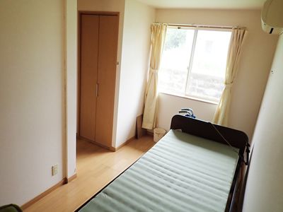 大きな窓にはカーテンが付き、近くにごみ箱が置かれ、壁にはエアコン、壁際には介護ベッドがあり、備え付けのクローゼットもある。