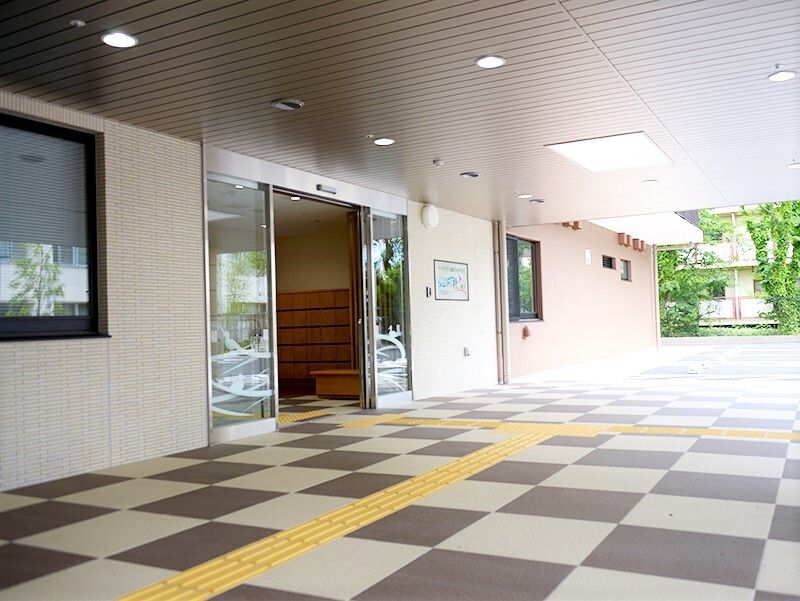 大きなガラス戸の自動ドアが設置されたエントランスには何も置かれていない。白とグレーのモザイク柄のフロアには点字ブロックが敷かれている。