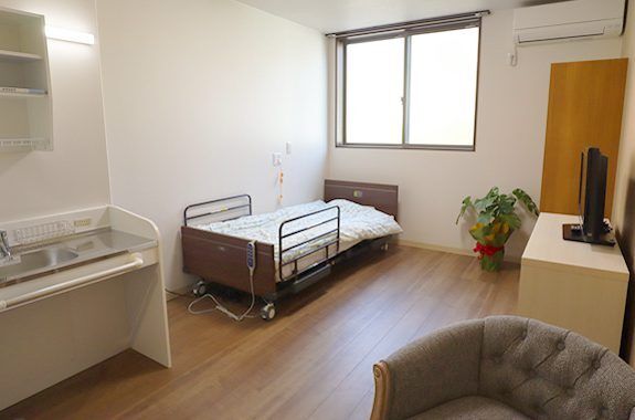 大きな窓が付いたフローリングの部屋。流し台やエアコン、一人用のソファ、ベッドがあり、ベッドサイドには緊急通報装置が付いている。