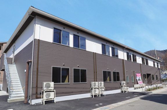 2階部分が白、1階部分がダークブラウンで三角屋根が付き、横に長い建物。側面には屋外階段があり、窓もたくさんついている。