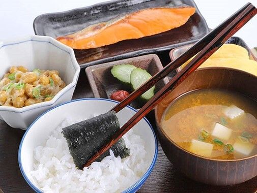 白いご飯に箸で海苔を巻いている。みそ汁や焼き鮭、出汁巻き卵、納豆などが並べられたトレー。