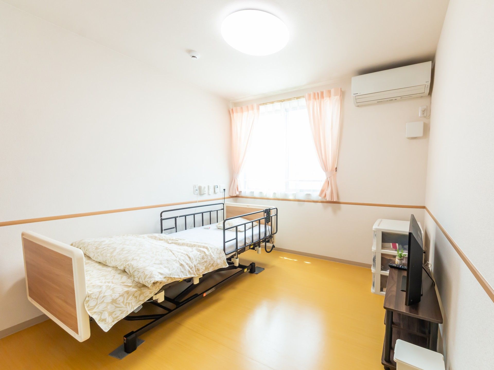 レースカーテンとカーテンが付いた大きな窓が二つ付いた部屋には寝具付きのベッドや車いすがあり、テレビも置かれている。