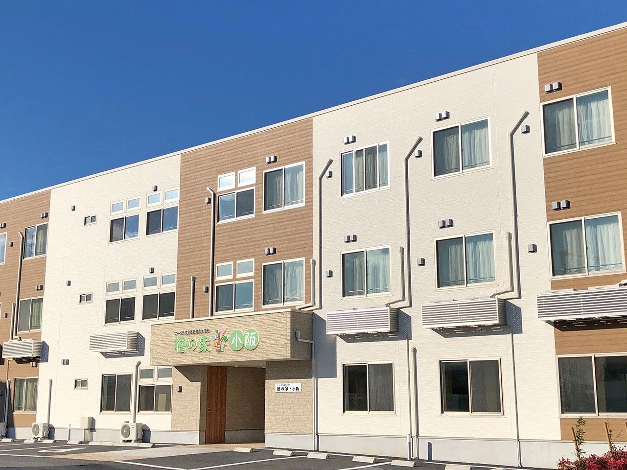 ベージュとブラウンの3階建ての建物。住宅名が書かれた屋根が玄関に付き、白線が引かれた駐車場もある。
