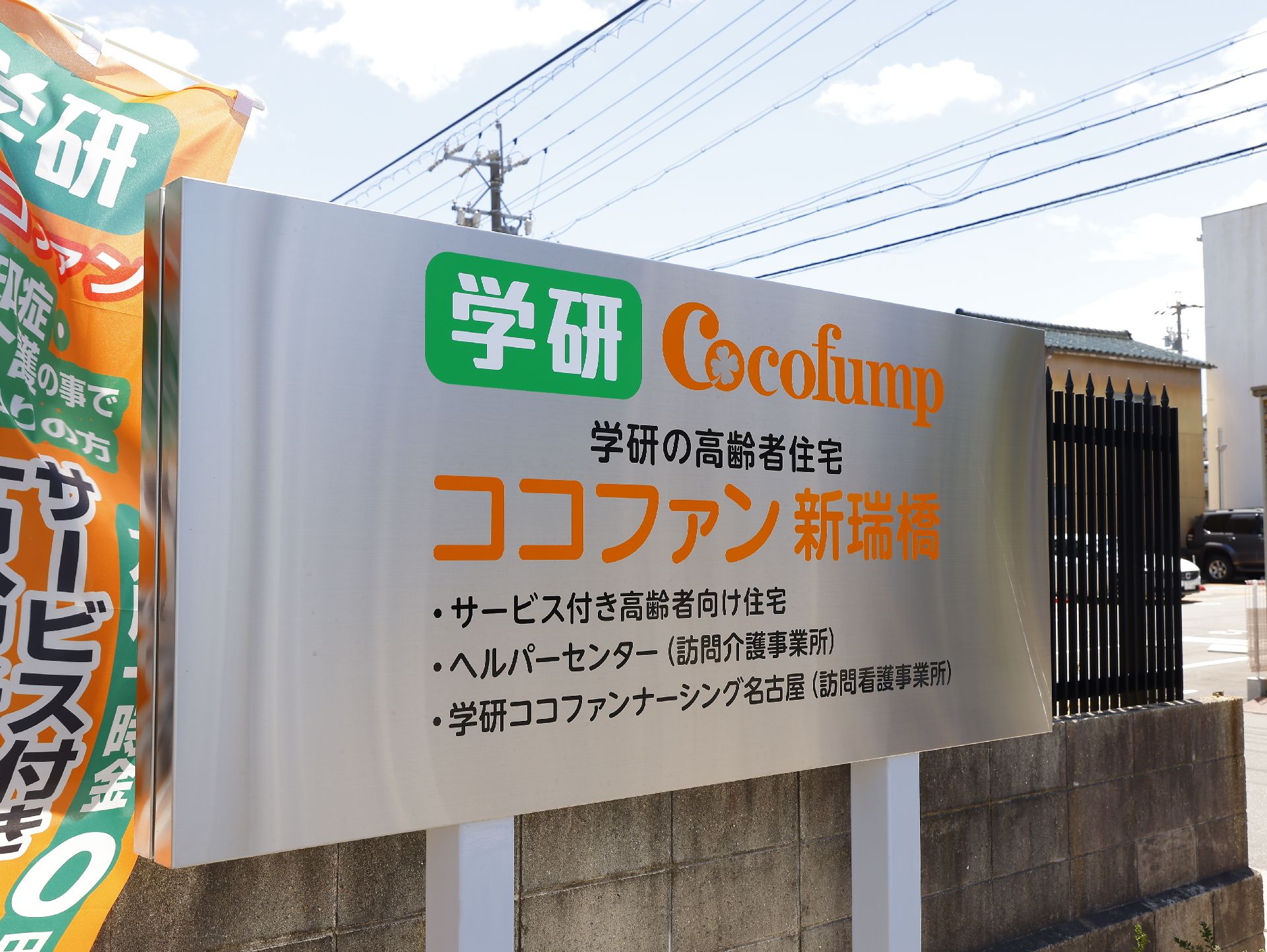 車いすの男性とピンクのエプロンを付けた女性スタッフが屋外で散策を楽しんでいる様子。周辺には緑が多く、二人は笑顔で過ごしている。