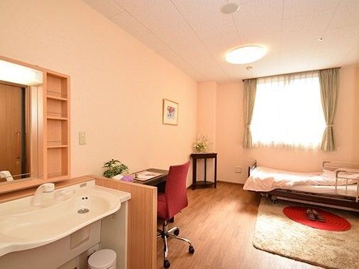 車いす対応のワイドタイプの洗面所が付いたフローリングの居室。大きな窓のそばには寝具付きのベッドがあり、デスクセットなども置かれている。